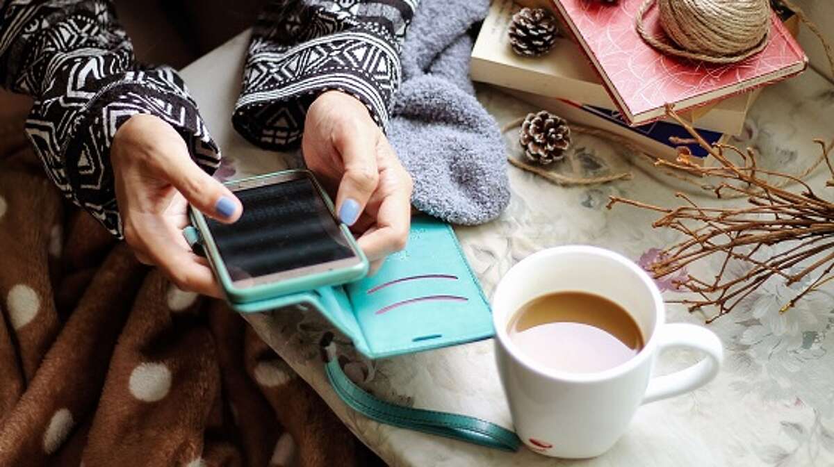 Hände, die, an einen Tisch gelehnt, ein Smartphone halten. Im Vordergrund ist eine Kaffeetasse zu sehen.