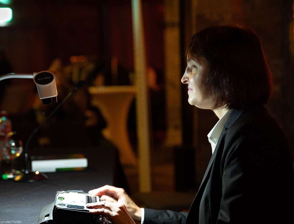 Die Lesende Sabine Lohner im Profil im abgedunkelten Raum, Lichtkegel auf den Händen, die auf der elektronischen Braille-Zeile liegen.